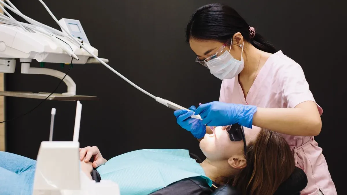 Dental professional in pink scrubs, mask and safety glasses cleaning a reclining patient's teeth with a corded dental instrument