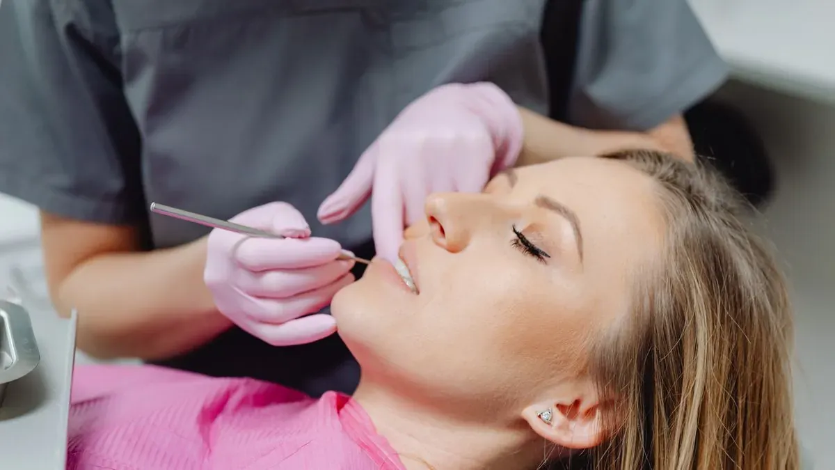 A dentist in pink gloves examining a young woman's teeth with a dental instrument