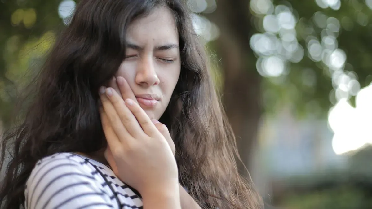 A young woman in a striped shirt pressing a hand to her cheek with a pained expression
