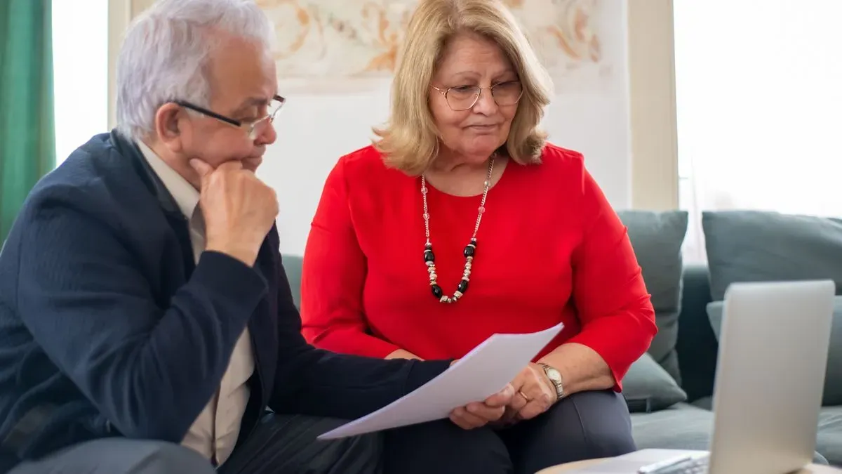 An older couple on a sofa reading a document together, with a laptop on the table