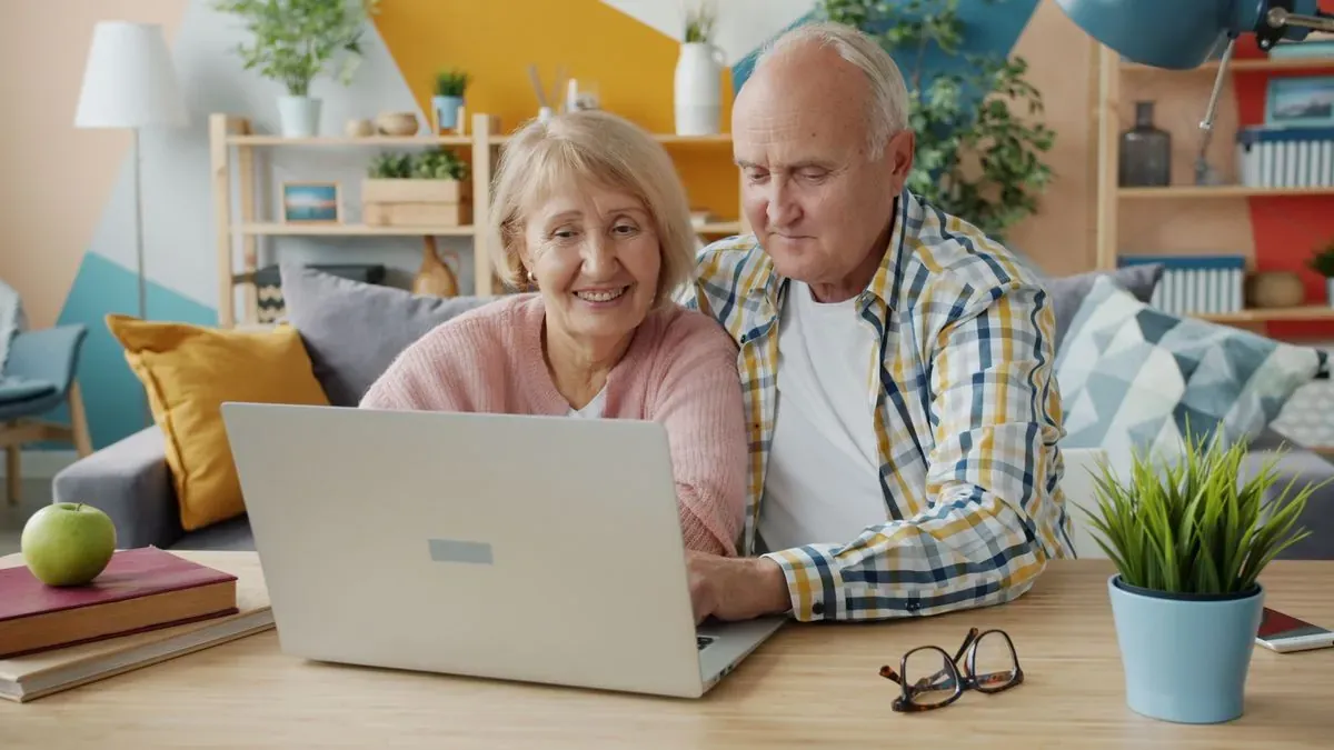 An older couple sitting at a table and looking at a laptop together in a bright living room