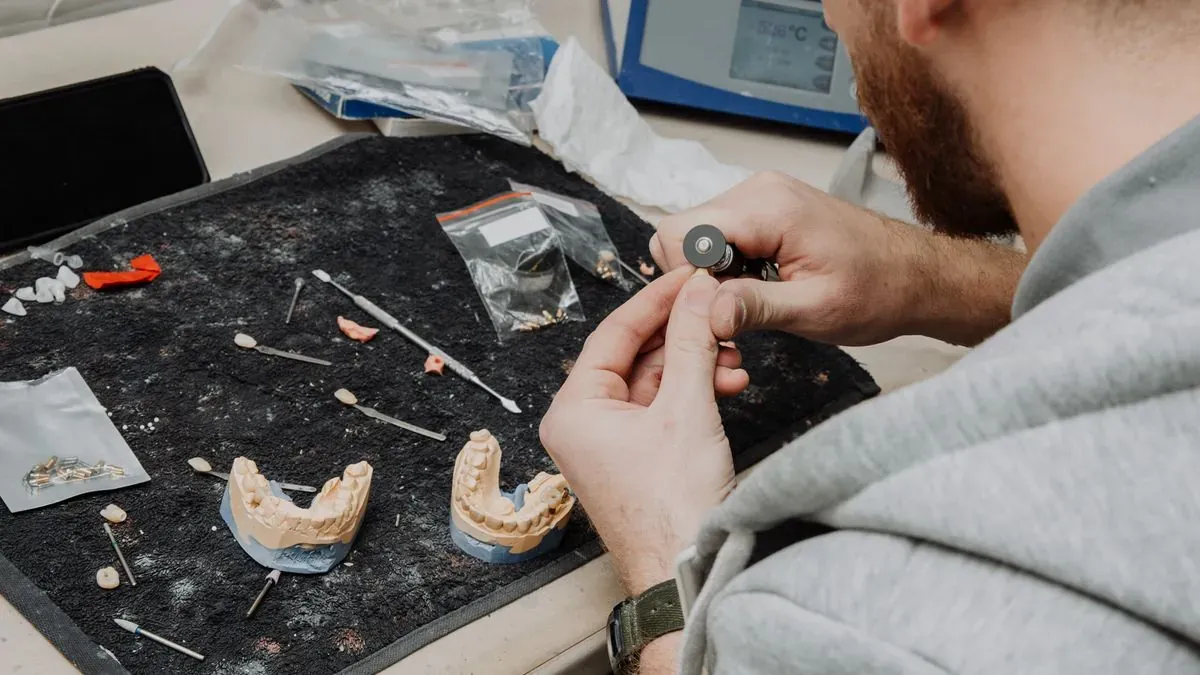 A dental technician's hands working at a table beside two plaster dental casts and small tools