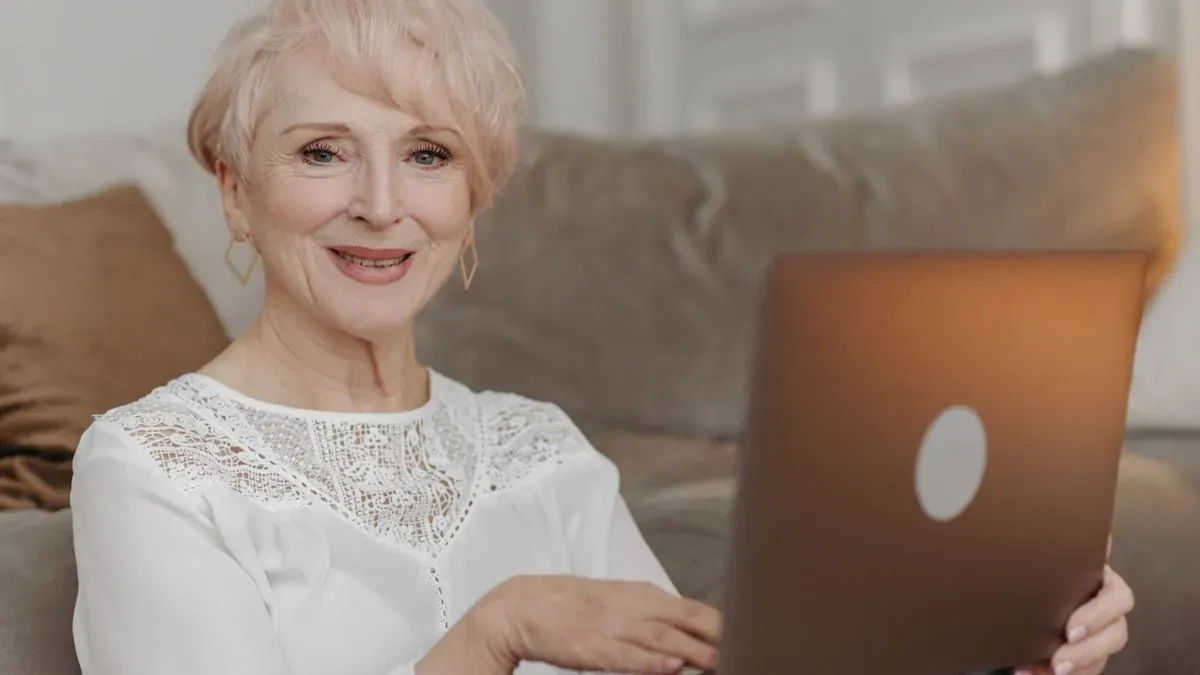 A smiling older woman with short blonde hair sitting on a sofa with a laptop on her lap