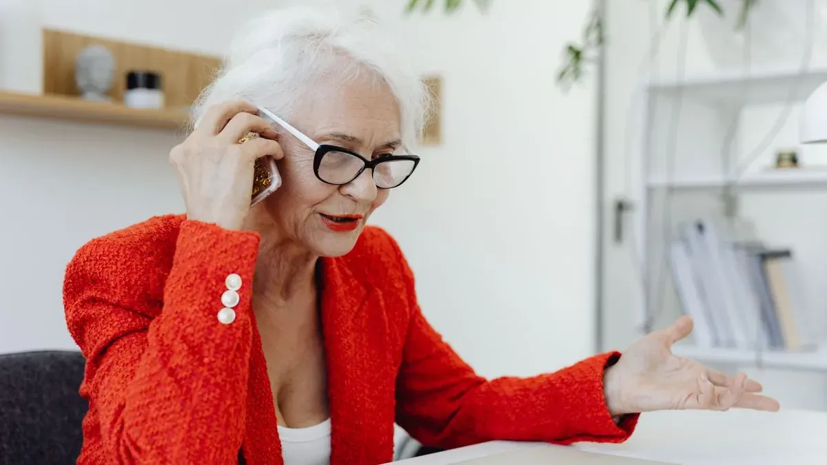 An older woman with white hair and glasses talking on the phone at a desk with a laptop
