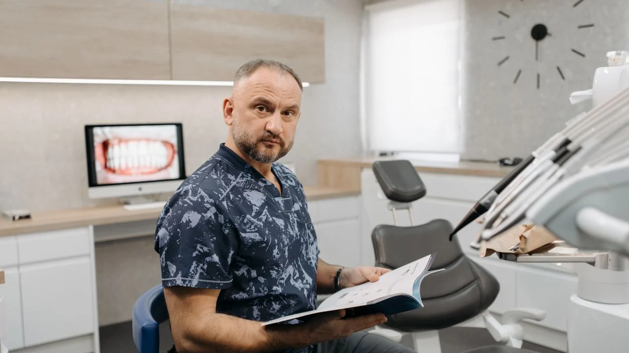 Dental Insurance for Self-Employed — Low Premium vs High Coverage Dentist in blue scrubs sitting in a dental office with dental chair and X-ray on monitor — comparing low-premium vs high-coverage dental insurance for self-employed workers