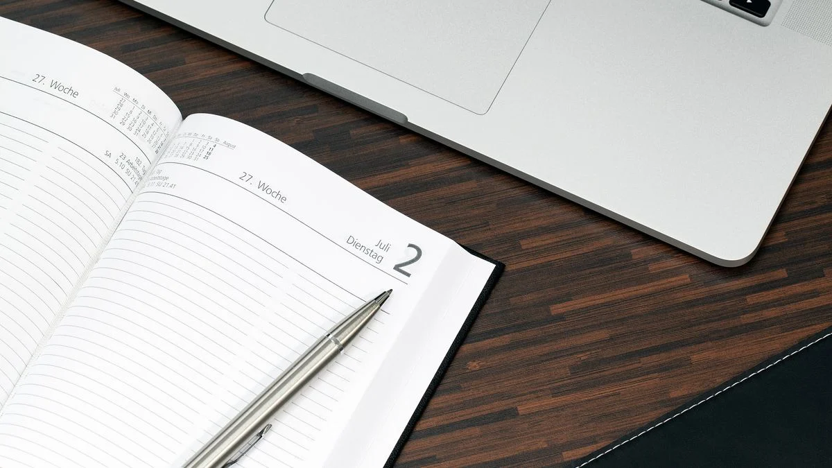 Open planner with a pen next to a laptop on a wooden desk, used for planning when to enrol in dental coverage