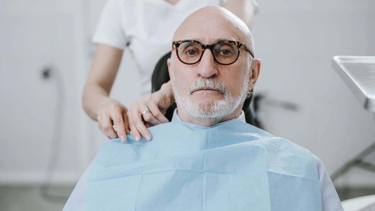 Older man in a dental chair being treated by a dentist, representing the crowns and dentures that carry a twelve-month wait