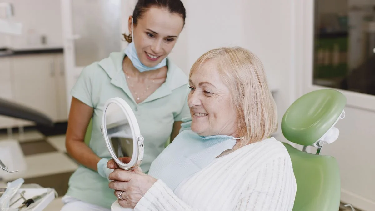 Senior woman checking her teeth in a mirror with her dentist Senior woman smiles while checking her teeth in a mirror with a dentist during a dental implant consultation.