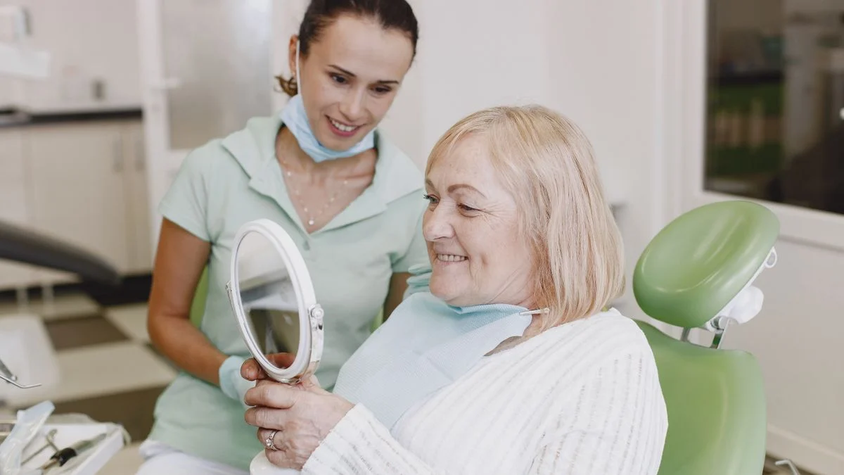 A senior woman smiles while checking her teeth in a mirror with a dentist, reflecting a successful, confirmed appointment.