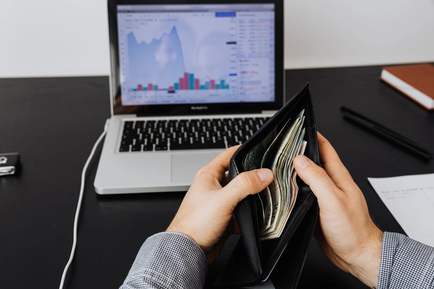A person holding a wallet with cash in front of a laptop showing financial charts