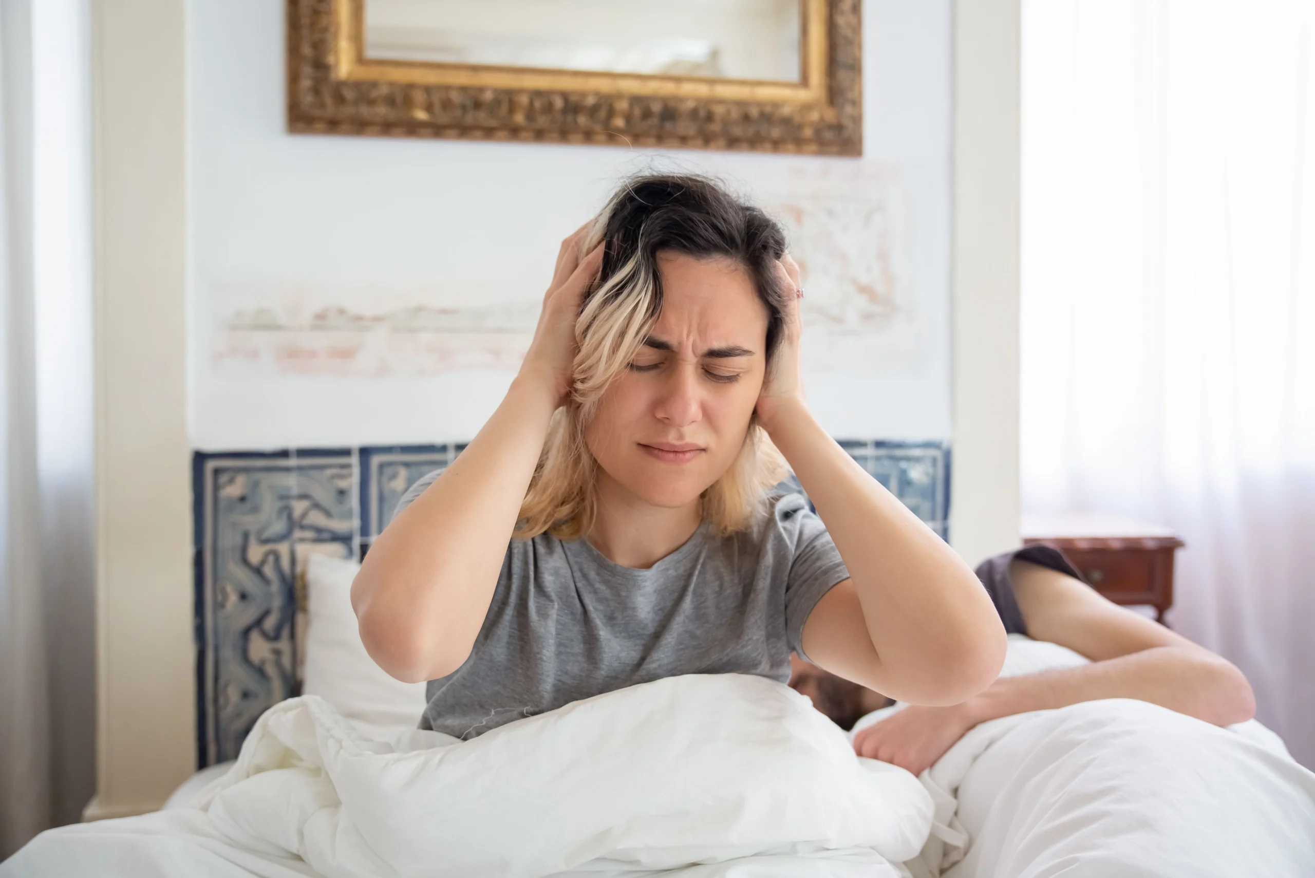 Waking with jaw tension from night-time clenching A woman sitting on a bed in a sunlit room holding her head, showing morning jaw and temple discomfort
